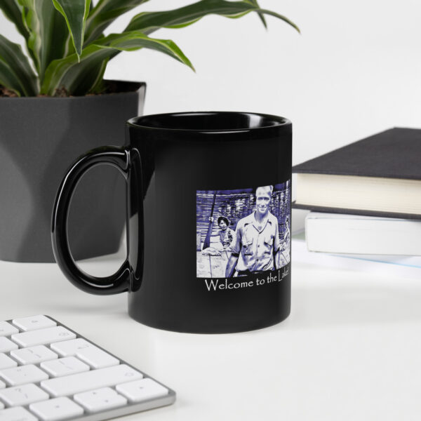 black glossy mug black 11 oz handle on left with image of the book cover for 'The History of Silver Lake as Told Through Its Deeds' showing an elderly man, Pappy, in foreground, two young women in a canoe behind him, and a young girl peeking from behind a tree saying ‘Welcome to the Lake’ on the historyofsilverlake.com website.