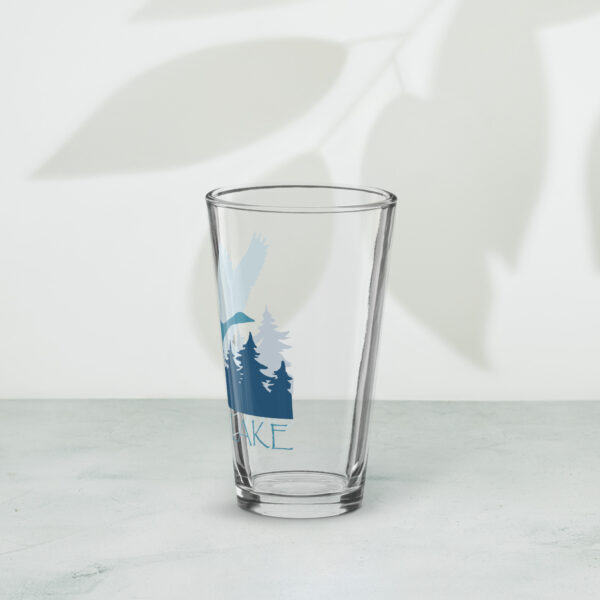 shaker pint glass 16 oz 16 oz left with a stylized teal blue logo for 'History of Silver Lake' book: A single goose flying over Silver Lake with trees in the background.